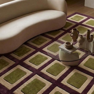 May include: A curved beige sofa sits on a rectangular area rug featuring a grid pattern of deep purple lines, light beige squares, and olive green rectangles. A glass coffee table holds several beige ceramic vases and a small book.