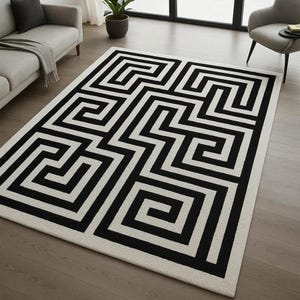 May include: A rectangular area rug with a geometric black and white pattern. The rug features a repeating maze-like design in black against a white background. The rug is placed on a hardwood floor in a living room setting.
