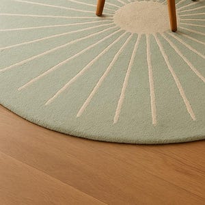 May include: A round, light green rug with a sunburst design. The center is a lighter shade of green, and the rays are off-white. The rug is partially visible, with a wooden chair leg in the frame. The rug is on a wooden floor.