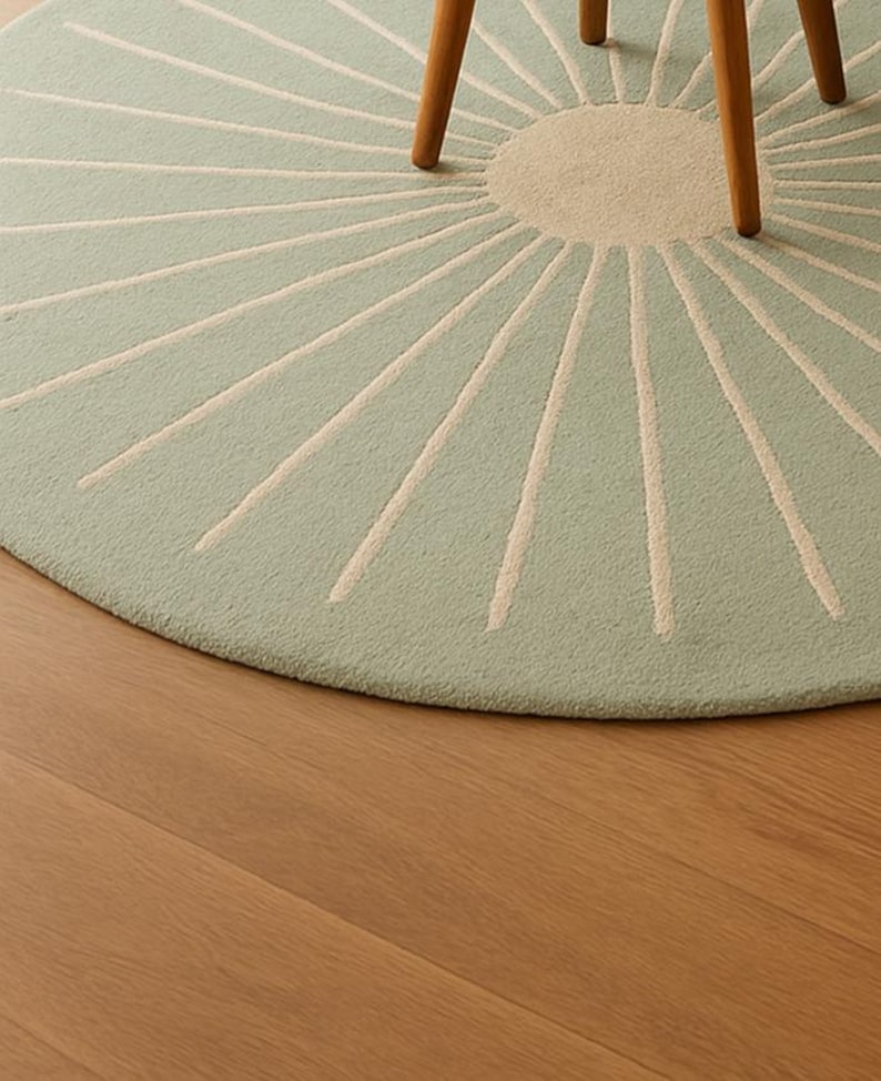 May include: A round, light green rug with a sunburst design. The center is a lighter shade of green, and the rays are off-white. The rug is partially visible, with a wooden chair leg in the frame. The rug is on a wooden floor.