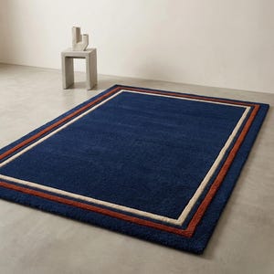 May include: A rectangular navy blue rug with a bordered design. The border features a rust-colored stripe, a cream-colored stripe, and a navy blue edge. The rug is placed on a concrete floor.