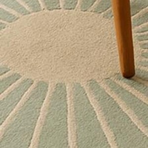 May include: Close-up of a round, light blue and beige rug with a sunburst design. The rug has a textured surface and is partially obscured by a wooden chair leg.