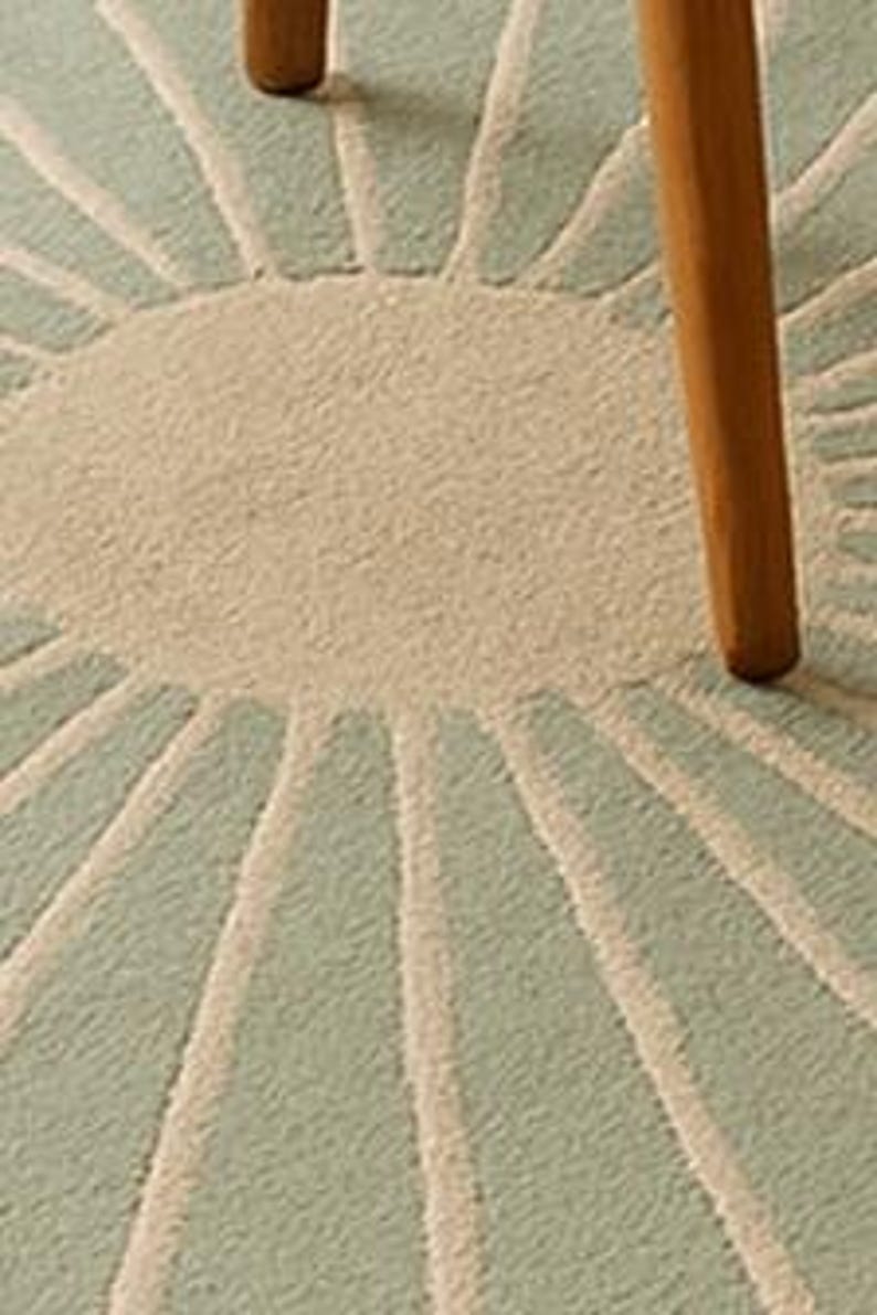 May include: Close-up of a round, light blue and beige rug with a sunburst design. The rug has a textured surface and is partially obscured by a wooden chair leg.