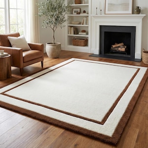 May include: A rectangular area rug featuring a large cream-colored center panel framed by a thick, dark brown border. The rug has a soft, textured pile and is positioned flat on a wooden floor, showcasing a clean, simple geometric design.