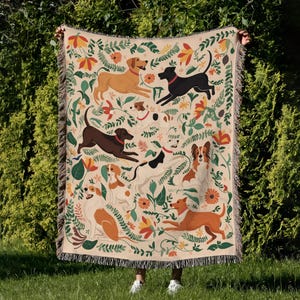 May include: A beige woven throw blanket with a pattern of dogs, flowers, and leaves. The dogs are in brown, black, and white, with red collars. The blanket has a fringed edge. The design includes various dog breeds and floral elements.