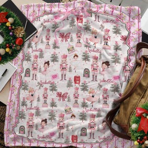 May include: A pink and white blanket featuring a pattern of nutcrackers, ballerinas, Christmas trees, and bows. The name "Isabella" is repeated throughout the design. The blanket has a scalloped edge and a pink ribbon tied at the top.