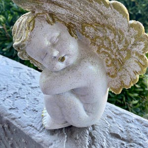 Puede incluir: Pequeña figura de querubín blanco con los ojos cerrados, sentado con las rodillas recogidas. El querubín tiene alas y detalles dorados. La figura descansa sobre una superficie gris, con follaje verde al fondo.