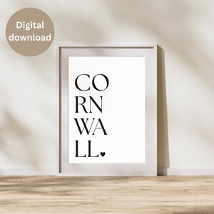 May include: White framed print with the word "CORNWALL" in black, vertically stacked text. A small black heart is at the end of the word. A digital download label is in the upper left corner. The print is on a white background.