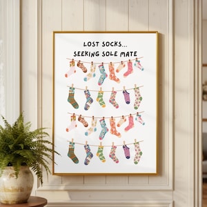 Op de afbeelding: Een ingelijste wandkunstprint met de tekst LOST SOCKS... SEEKING SOLE MATE boven vier horizontale waslijnen. Elke lijn toont een verscheidenheid aan kleurrijke, bedrukte sokken die met houten wasknijpers aan een witte achtergrond hangen.