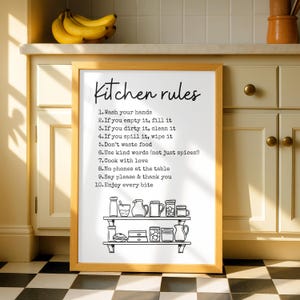 Puede incluir: Un marco de madera con las palabras "Kitchen rules" en una fuente manuscrita. La impresión enumera diez reglas para la cocina, incluyendo lavarse las manos, limpiar y disfrutar cada bocado. Una estantería con utensilios de cocina está en la parte inferior.