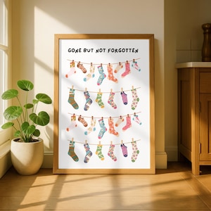 Peut inclure: Une affiche encadrée avec le texte GONE BUT NOT FORGOTTEN en haut, présentant quatre cordes à linge horizontales avec diverses chaussettes colorées et à motifs suspendues par des pinces à linge en bois sur un fond blanc.