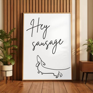 Pode incluir: Impressão de arte emoldurada com as palavras "Hey sausage" em escrita preta, sobre um desenho minimalista de um dachshund. A obra de arte é exibida em um fundo branco com uma moldura de madeira.