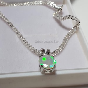 Könnte beinhalten: Eine silberne Halskette mit einem Anhänger aus weißem Opal. Der Opal zeigt grüne und andere Farbtöne. Die Kette hat ein filigranes, gegliedertes Design. Der Schmuck wird in einer weißen Schachtel präsentiert.