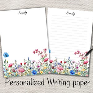 Peut inclure: Deux feuilles de papier à lettres personnalisées avec une bordure florale. L'une est vierge, l'autre lignée. Le nom "Emily" est en haut. Un stylo noir repose sur la feuille lignée. La bordure inférieure présente des fleurs colorées.
