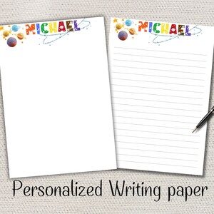 Peut inclure: Deux feuilles de papier à écrire blanches personnalisées avec le nom "MICHAEL" en lettres colorées. Le haut du papier présente des planètes et des étoiles. Une feuille est vierge, l'autre est lignée. Un stylo noir repose sur la feuille lignée. Le texte "Personalized Writing paper" est en bas.
