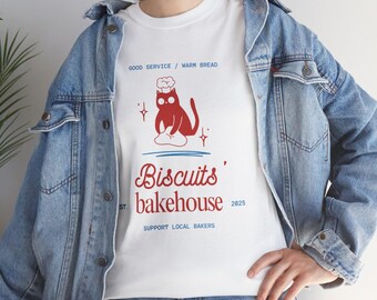 May include: White t-shirt with a red graphic of a cat wearing a chef's hat, sitting on a cloud. The text reads "Biscuits' Bakehouse" with "Good Service / Warm Bread" above and "Support Local Bakers" below.