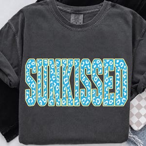 以下が含まれることがあります： チャコールグレーの半袖Tシャツで、カレッジスタイルの太字フォントで「SUNKISSED」という文字がプリントされています。文字は鮮やかなブルーのヒョウ柄で塗りつぶされており、薄いライトイエローの縁取りが施されています。