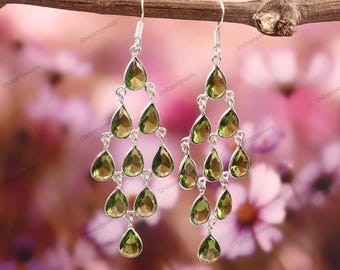 May include: Silver chandelier earrings with multiple pear-shaped, olive-green gemstones. The earrings are suspended from a branch against a soft pink and purple floral background. The earrings are labeled with the text "Shibamjewels".
