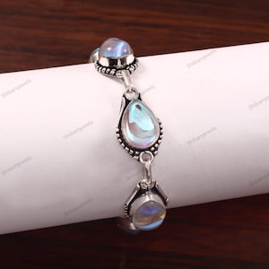 Op de afbeelding: Een zilveren armband met drie iriserende maansteen cabochons. De armband heeft een sierlijk ontwerp met een druppelvormige steen in het midden en ronde stenen aan elk uiteinde. De stenen reflecteren tinten blauw en wit.
