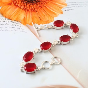 Op de afbeelding: Een zilveren armband met ovale, dieprode edelstenen. De armband bestaat uit meerdere edelstenen die met zilveren schakels zijn verbonden, met kleine bloemmotieven tussen sommige stenen. De sluiting is een eenvoudige haak- en oogsluiting.