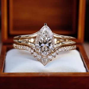 Puede incluir: Un juego de anillos de compromiso de oro con un diamante central en forma de pera. El anillo va acompañado de una banda con diamantes y una banda curva, todo en una caja de anillos de madera. El diamante es claro y brillante.