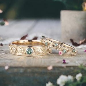 May include: Two gold rings with leaf and vine detailing. One ring features a central teal gemstone, while the other has multiple smaller teal gemstones. The rings are set on a wooden surface.