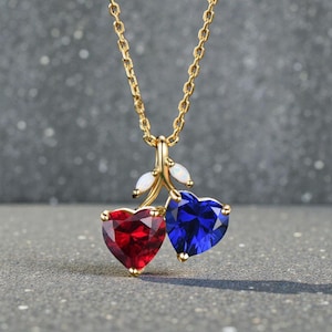 May include: A gold necklace with a cherry pendant. The pendant has two heart-shaped gemstones, one red and one blue, with white accents. The chain is a delicate gold link. The gemstones are in the shape of hearts.