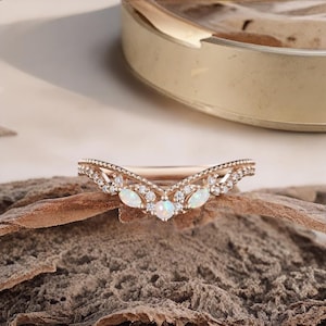 May include: A rose gold ring with a curved design, featuring small clear stones and three opal-like stones. The ring is set on a textured, brown surface, with a gold-coloured dish in the background.