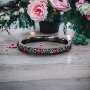 Könnte beinhalten: Ein schwarzer Ring mit roten Edelsteinen, die in einem dekorativen Muster gefasst sind. Der Ring liegt auf einer Holzoberfläche, mit verstreuten rosa Blütenblättern und einem verschwommenen Blumengesteck im Hintergrund. Das Design des Rings zeigt Wirbel und Blätter.