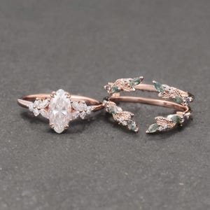 May include: Two rose gold rings. One ring features a marquise-cut diamond center stone, surrounded by smaller diamonds and floral accents. The second ring has a leaf design with green and clear stones.
