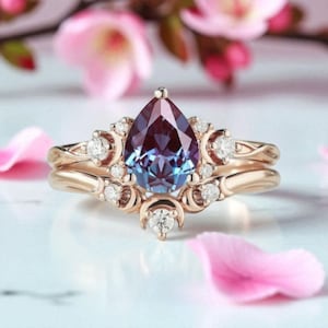 May include: A gold-colored ring set featuring a pear-shaped, color-changing gemstone. The ring is accented with small, round, clear gemstones and decorative swirls. The background includes pink flower petals and soft focus floral elements.