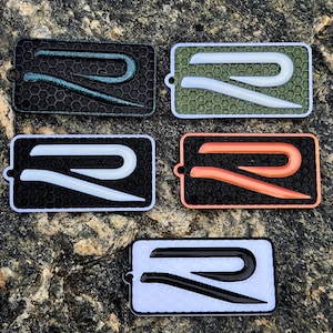 May include: Five rectangular patches featuring a raised letter R design over a honeycomb texture. The patches come in various colour combinations: black with teal, olive green with white, black with white, black with orange, and white with black.