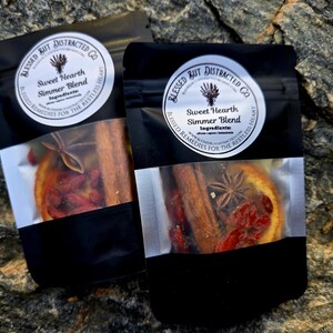 May include: Two black pouches with clear windows displaying a simmer blend. Each pouch has a white label with text that reads "Sweet Hearth Simmer Blend". The blend includes cinnamon sticks, star anise, and dried orange slices.