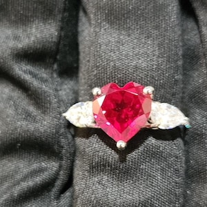 Può includere: Un anello d'argento con una grande pietra preziosa rossa a forma di cuore come elemento centrale. Due pietre preziose chiare più piccole a forma di pera affiancano il cuore. L'anello è mostrato su uno sfondo scuro.