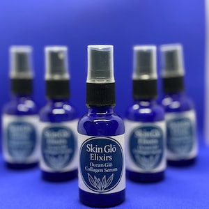 May include: A group of five cobalt blue glass bottles with black spray nozzles. Each bottle has a white label with the text "Skin Glo Elixirs Ocean Glo Collagen Serum". The background is a solid blue.