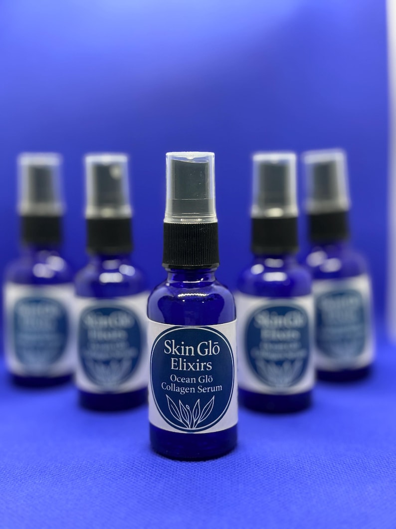 May include: A group of five cobalt blue glass bottles with black spray nozzles. Each bottle has a white label with the text "Skin Glo Elixirs Ocean Glo Collagen Serum". The background is a solid blue.