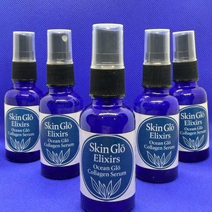 May include: Five cobalt blue glass bottles of Skin Glo Elixirs Ocean Glo Collagen Serum. Each bottle has a black spray top and a white label with the product name and a botanical illustration. The bottles are arranged on a blue surface.