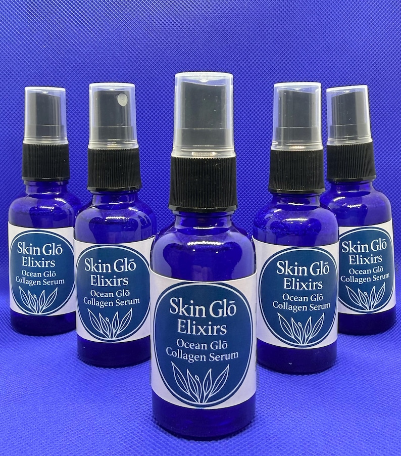 May include: Five cobalt blue glass bottles of Skin Glo Elixirs Ocean Glo Collagen Serum. Each bottle has a black spray top and a white label with the product name and a botanical illustration. The bottles are arranged on a blue surface.