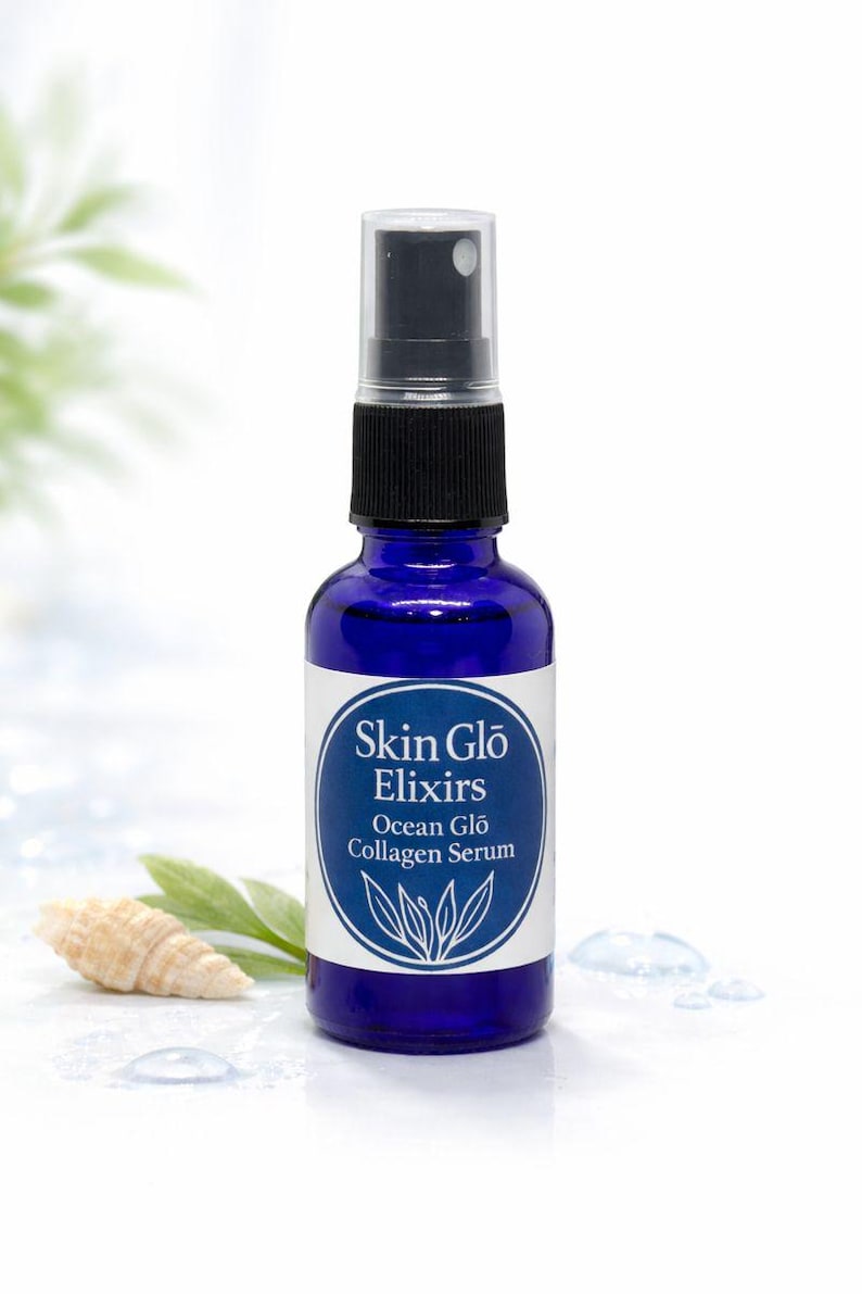 May include: A cobalt blue glass bottle of Skin Glo Elixirs Ocean Glo Collagen Serum. The bottle has a black spray nozzle and a white label with the product name and a botanical design. A seashell and leaves are in the background.