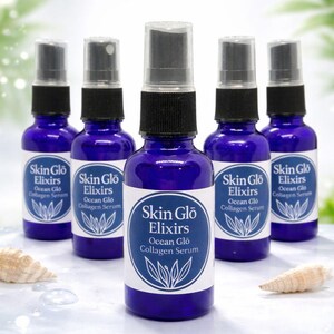 May include: Five cobalt blue glass bottles with black spray tops are arranged on a light blue surface. Each bottle has a white label with the text "Skin Glo Elixirs Ocean Glo Collagen Serum" and a botanical illustration. Seashells are in the foreground.