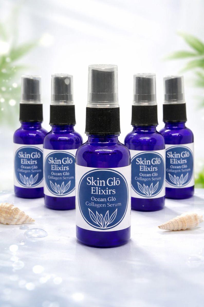 May include: Five cobalt blue glass bottles with black spray tops are arranged on a light blue surface. Each bottle has a white label with the text "Skin Glo Elixirs Ocean Glo Collagen Serum" and a botanical illustration. Seashells are in the foreground.
