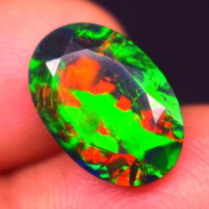 May include: An oval-shaped gemstone with a vibrant display of color. The gem features a mix of bright green, fiery orange, and dark blue hues. The stone is held in a hand, showcasing its polished facets and internal play of color.