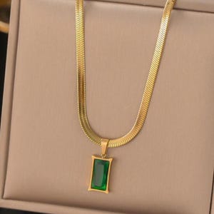 Puede incluir: Collar dorado con un colgante rectangular verde esmeralda. La cadena tiene un diseño plano en espiga. El colgante está enmarcado en oro y presenta una gema verde vibrante. El collar se muestra sobre un fondo neutro.