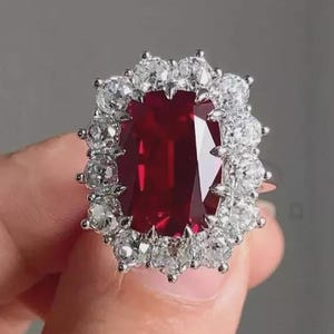 Könnte beinhalten: Ein auffälliger Ring mit einem großen, rechteckigen, tiefroten Edelstein, umgeben von einem Halo aus kleineren, funkelnden weißen Steinen. Die Fassung des Rings ist silberfarben und bildet einen auffälligen Kontrast.