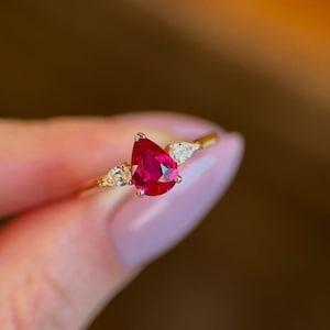 May include: A gold ring with a striking, pear-cut, deep red gemstone at its centre. The ring is embellished with smaller, clear gemstones on either side of the main stone. The ring's band is a delicate gold colour, creating an elegant and eye-catching piece of jewellery.