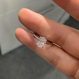 May include: A silver ring featuring a large, oval-cut diamond. The diamond is the central element, catching the light. The band is slender and elegant, enhancing the gemstone's size. The ring is held between fingers, highlighting its design.