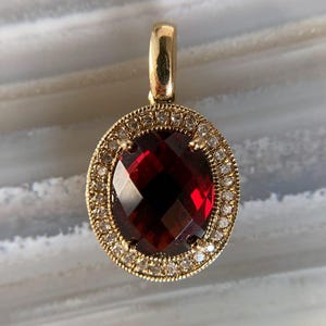 Könnte beinhalten: Ein goldener Anhänger mit einem großen, facettierten, tiefroten Edelstein in der Mitte. Der Edelstein ist von einem Kranz aus kleinen, klaren Edelsteinen umgeben. Der Anhänger hat ein schlichtes, elegantes Design, für eine Halskette.