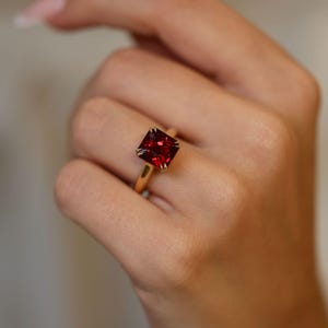 May include: A gold ring with a large, square-cut, deep red gemstone. The gemstone is set with four prongs, and the band is a simple, polished gold. The ring is shown on a hand.