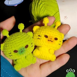 Op de afbeelding: Twee handgemaakte gehaakte alien-speeltjes, één limoengroen en één geel, worden in een hand gehouden. De aliens hebben kleine antennes, zwarte ogen en lachende gezichten. Een cartoon UFO en een kikker staan op de achtergrond.