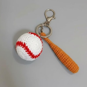 May include: A handmade baseball keychain featuring a white ball with red stitching and an orange crochet bat. The keychain has a silver clasp and ring. The baseball is approximately 3 inches in diameter.